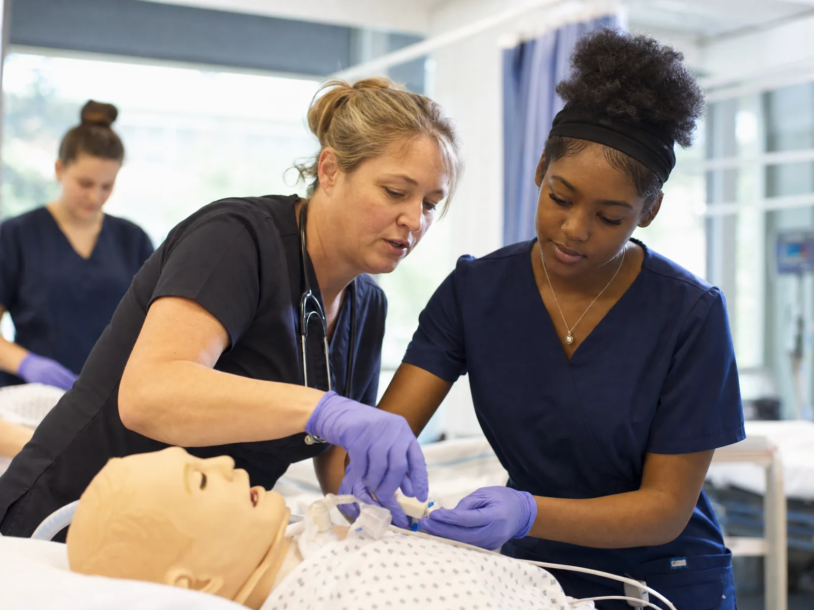 A nurse instructor guiding a CNA trainee through a hands-on procedure on a patient-care mannequin